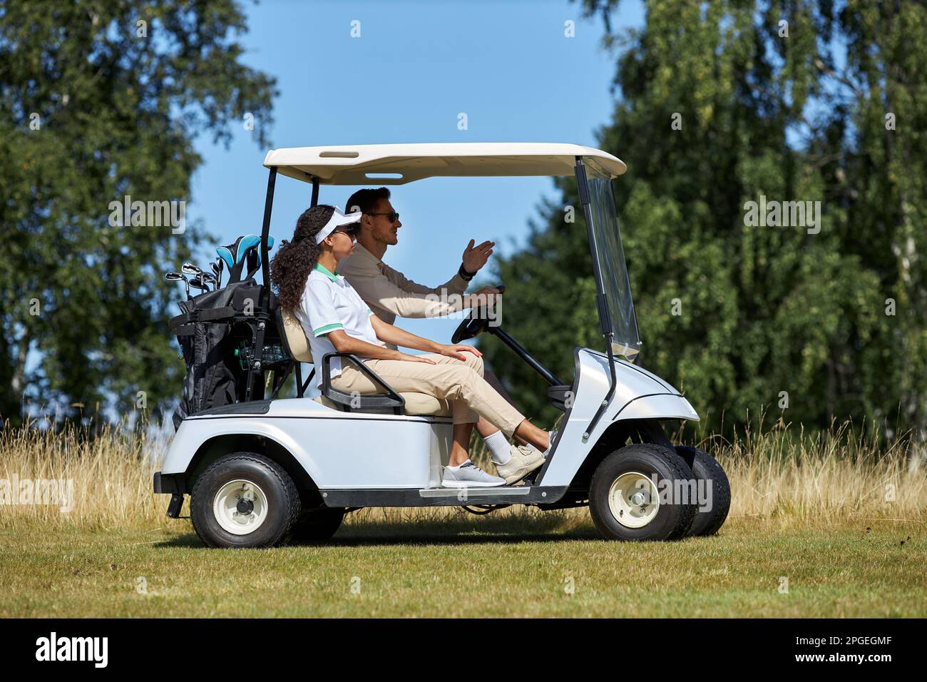 Seitliches Porträt eines sportlichen jungen Paares, das einen Golfwagen über das Feld im Sportclub fährt, der von Sonnenlicht beleuchtet ist Stockfoto