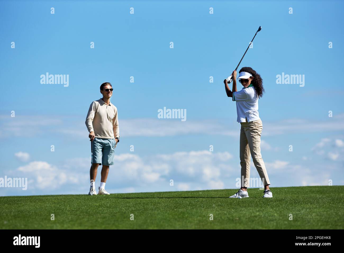 Ein langes Porträt eines aktiven, sportlichen Paares, das Golf gegen den blauen Himmel spielt, mit einer Frau, die Golfschläger spielt Stockfoto