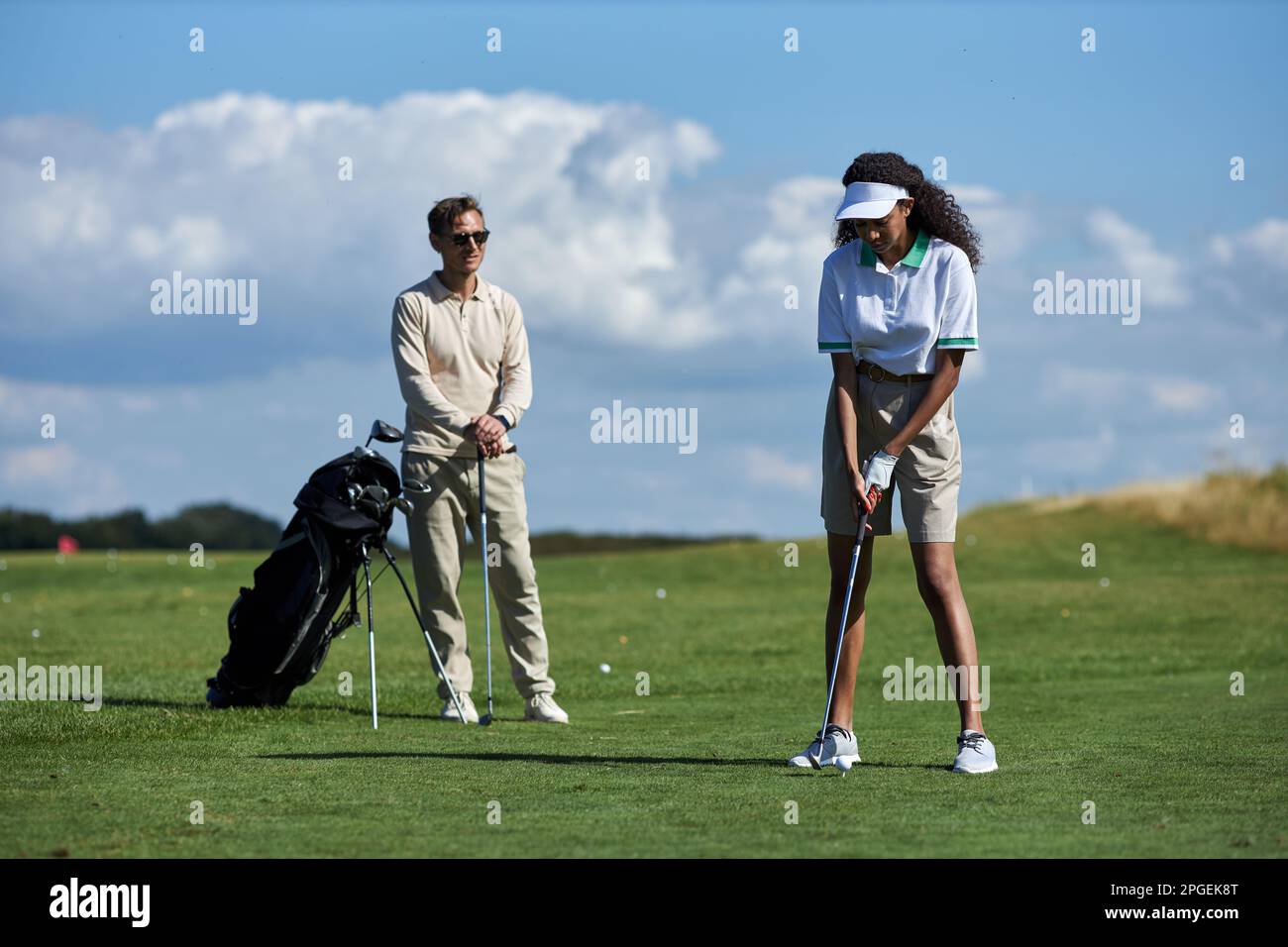 Ein langes Porträt eines sportlichen jungen Paares, das Golf auf dem grünen Feld spielt, mit Fokus auf einer schwarzen Frau, die mit dem Schläger Ball schlägt Stockfoto