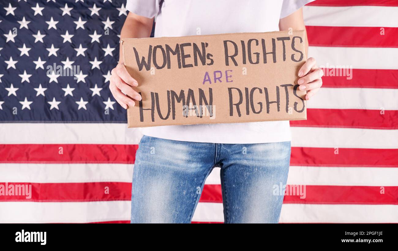 Junge Frau Protestierenden hält Pappe mit Womens Rights are Human Rights Zeichen gegen USA Flagge auf Hintergrund. Feministische Macht. Chancengleichheit für Frauen Stockfoto