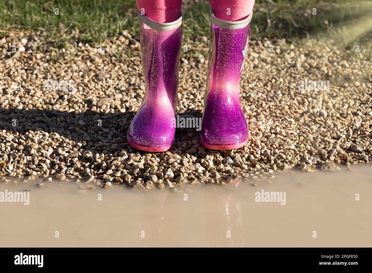 Ein Kind trägt neue, leuchtend pinkfarbene Gummistiefel, steht neben einer schlammigen Pfütze, kein Gesicht. Kindergartenkinder. Schuhe für Regenwetter Stockfoto