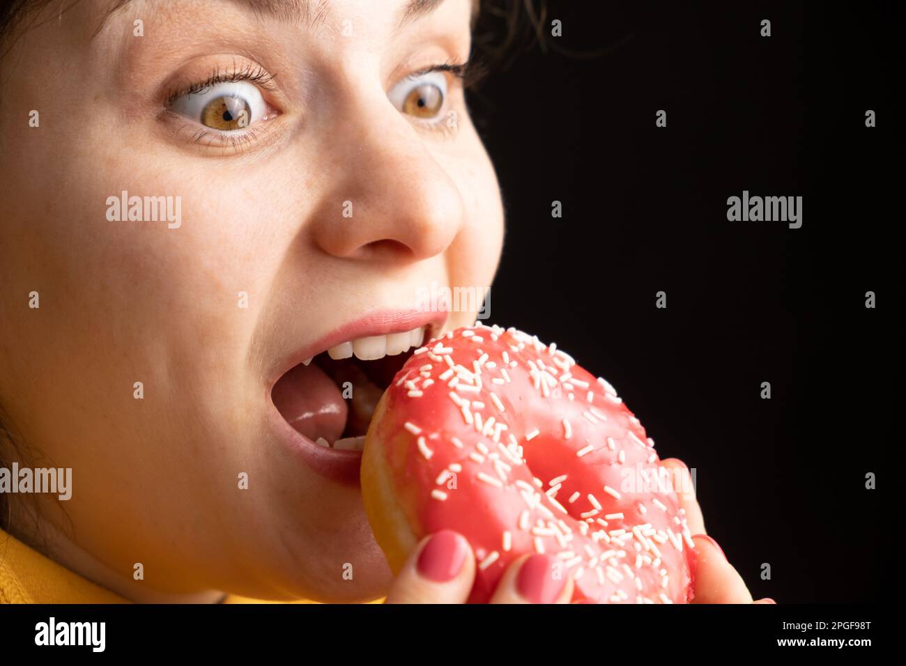 Eine Frau beißt einen großen roten Donut, einen schwarzen Hintergrund, ein Ort für Text. Gluttonie, überfressen und zuckersüchtig. Stockfoto