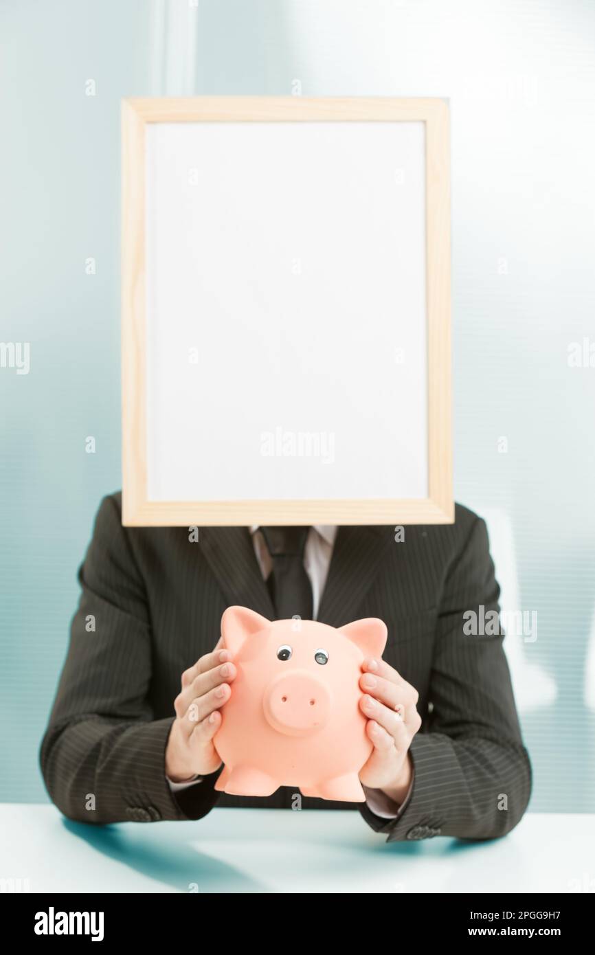 Büroarbeitsplatz, ein Mann mit Schildern, eingerahmt in hellem Holz, hat ein Sparschwein. Er hält sie für ein Konzept von Einsparungen, Ausgaben, Investitionen und Stockfoto