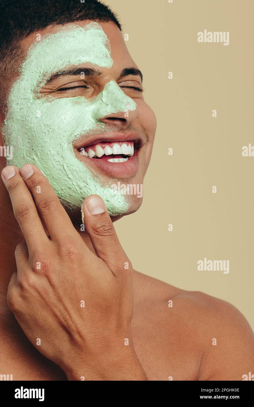 Ein junger Mann lächelt, während er sich mit einer grünen Gesichtsmaske um sich selbst kümmert. Ein glücklicher junger Mann, der seine Haut mit Wasser versorgt und verwöhnt Stockfoto