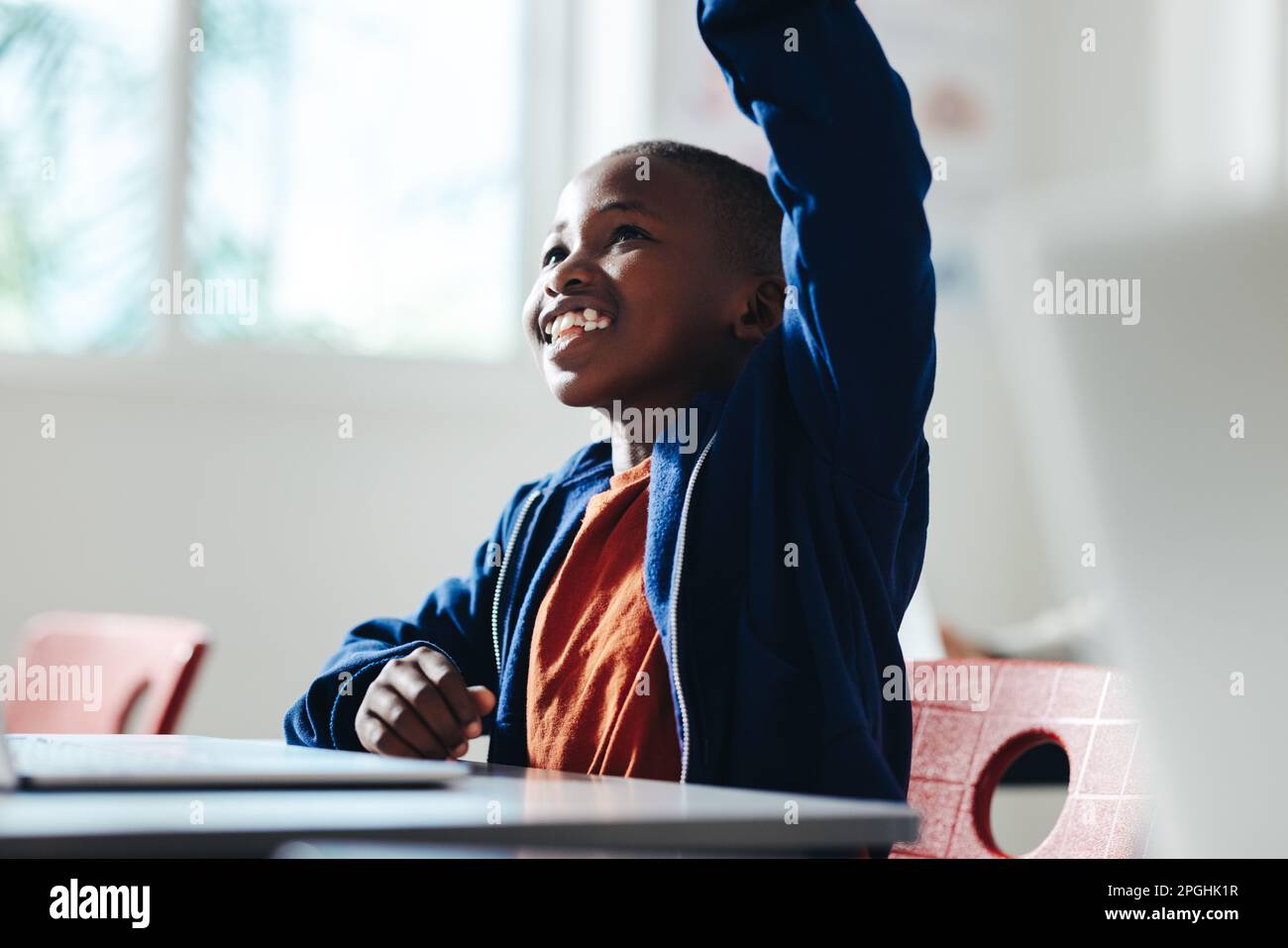 Ein schwarzer Junge, der in einer Grundschule die Hand hebt, um eine Frage zu beantworten. Mit einem selbstbewussten Lächeln im Gesicht, engagiert sich der Junge Stockfoto