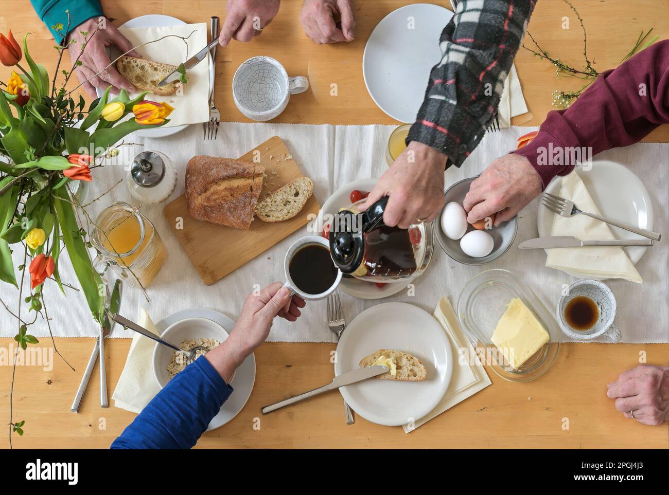 Blick von oben auf eine Gruppe von Personen, die auf einem Holztisch sitzen und zusammen frühstücken mit Kaffee, Brot, Eiern, Butter und einigen Frühlingsblumen, High A Stockfoto