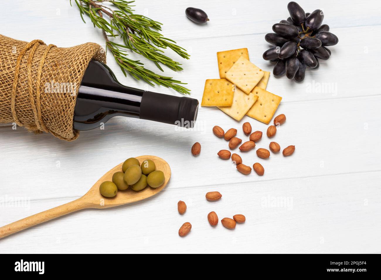 Grüne Oliven in Holzlöffel. Nüsse, Cracker und Zweige schwarzer Trauben auf dem Tisch. Eine Flasche Wein. Weißer Hintergrund. Stockfoto