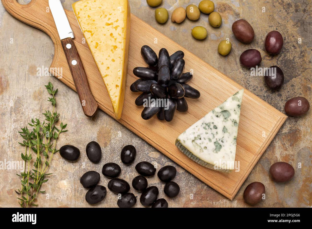 Blauschimmelkäse, Trauben und Messer auf dem Brett. Oliven und Thymianzweig auf dem Tisch. Rostiger Hintergrund. Stockfoto