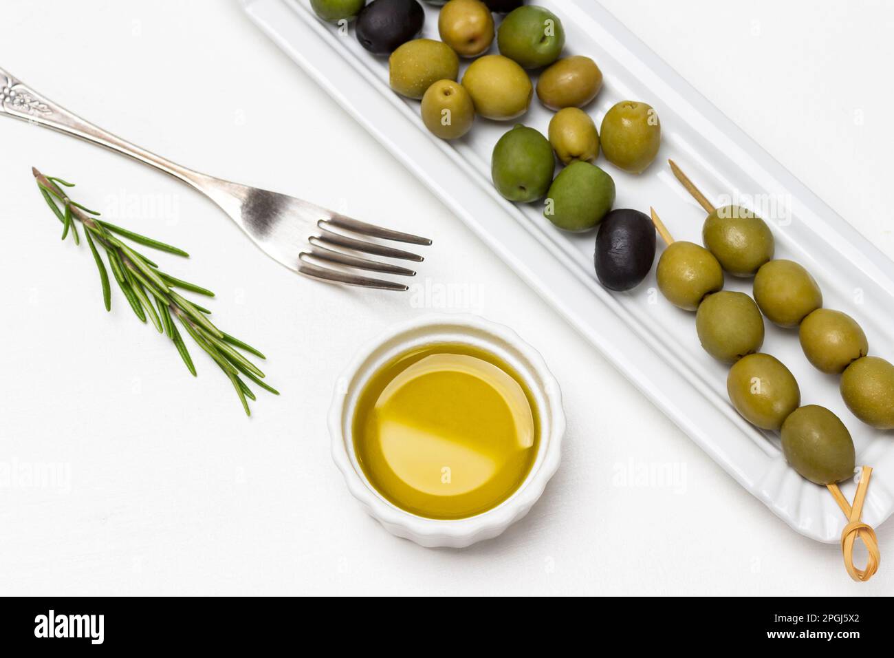 Grüne Oliven auf einem weißen Teller. Olivenöl in einer kleinen Schüssel. Gabel und Zweig Rosmarin auf dem Tisch. Weißer Hintergrund. Stockfoto