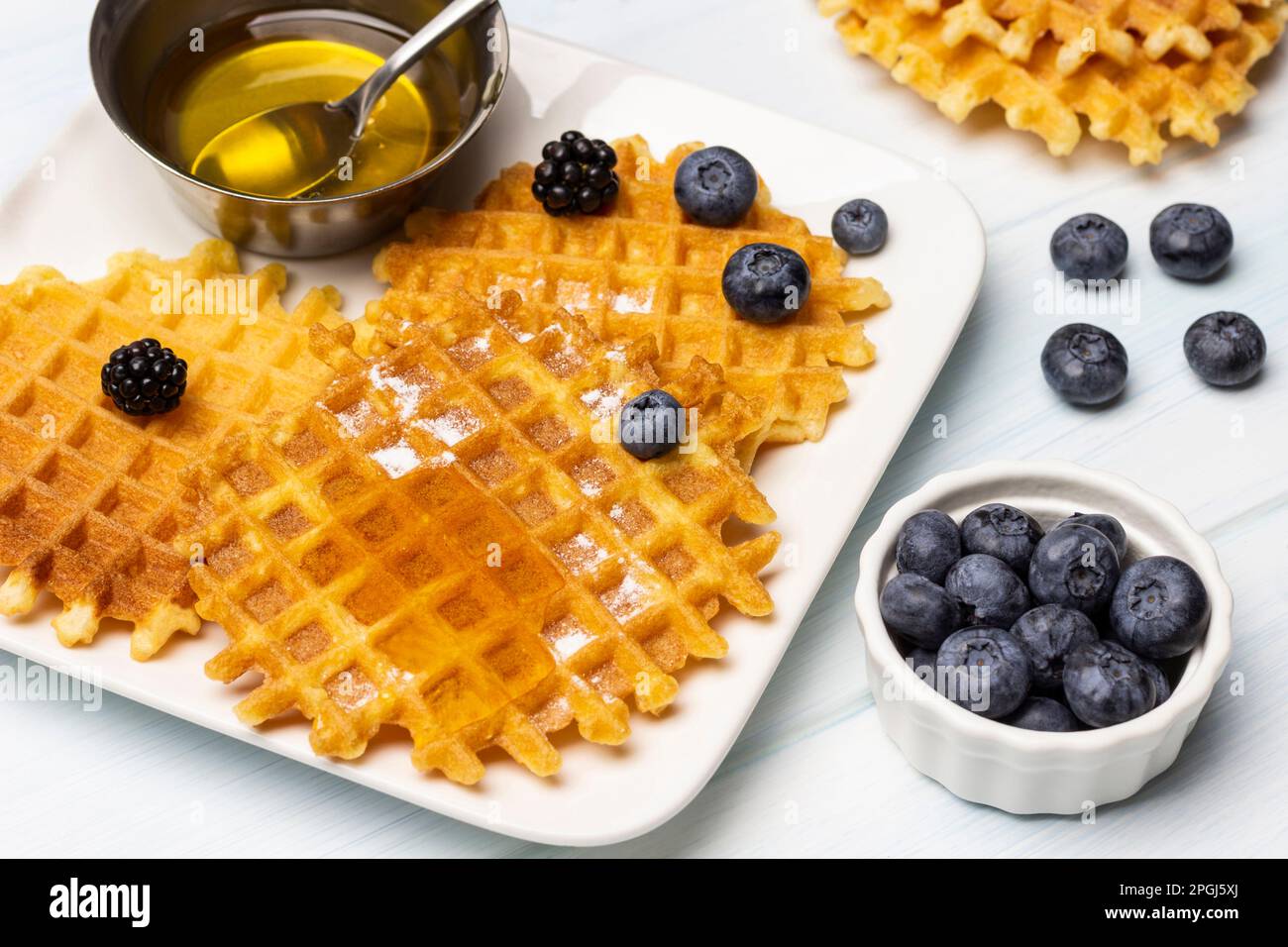 Waffeln und Beeren, ein Löffel in einer Metallschüssel mit Honig, auf einem weißen Teller. Blaubeeren in einer weißen Schüssel. Weißer Hintergrund. Stockfoto