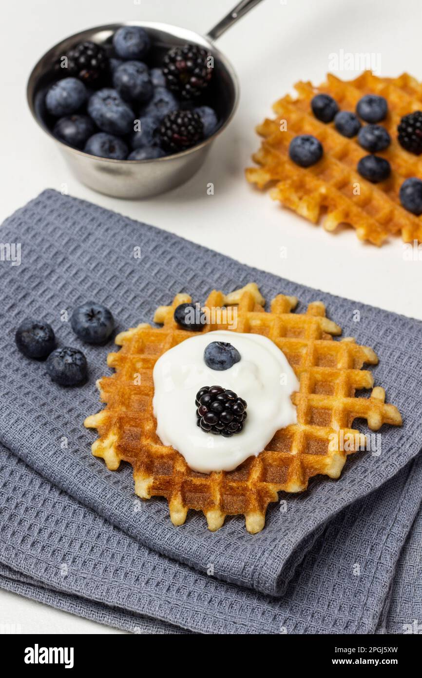 Beeren und Joghurt auf Waffeln auf grauer Serviette. Brombeeren und Blaubeeren in einer Metallschüssel auf dem Tisch. Weißer Hintergrund. Stockfoto