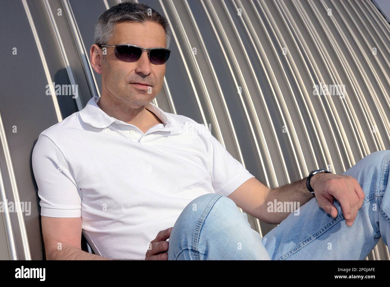 Ein älterer Mann mit Sonnenbrille und einem Poloshirt, der zwanglos vor einer Blechwand sitzt Stockfoto