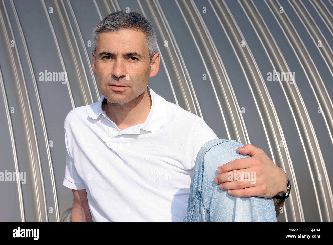 Ein älterer Mann in einem Poloshirt, der vor einer Blechwand sitzt Stockfoto