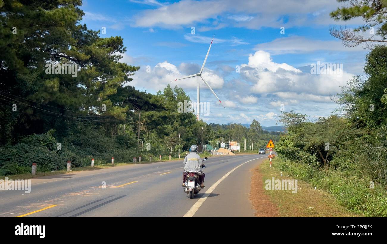 Ein Motorrad fährt die AH17. Straße in Vietnams Central Highlands in der Nähe von Buon Ma Thuot neben einer riesigen Windturbine. Stockfoto