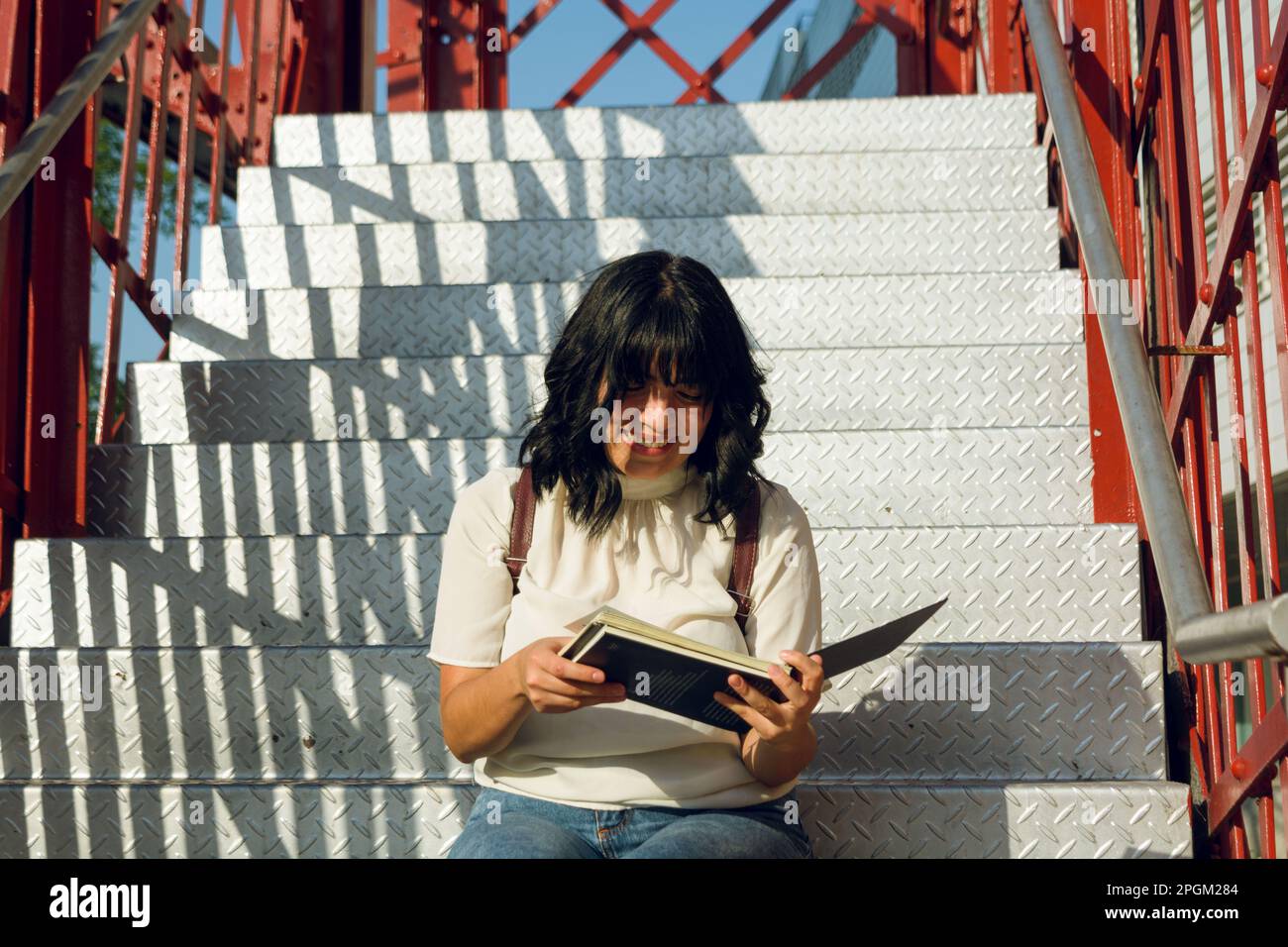 Vorbereitungs- und Universitätsstudium, junge lateinin, glücklich, ein Buch auf der Treppe zu lesen, sich darauf vorzubereiten, ein Profi zu werden. Stockfoto