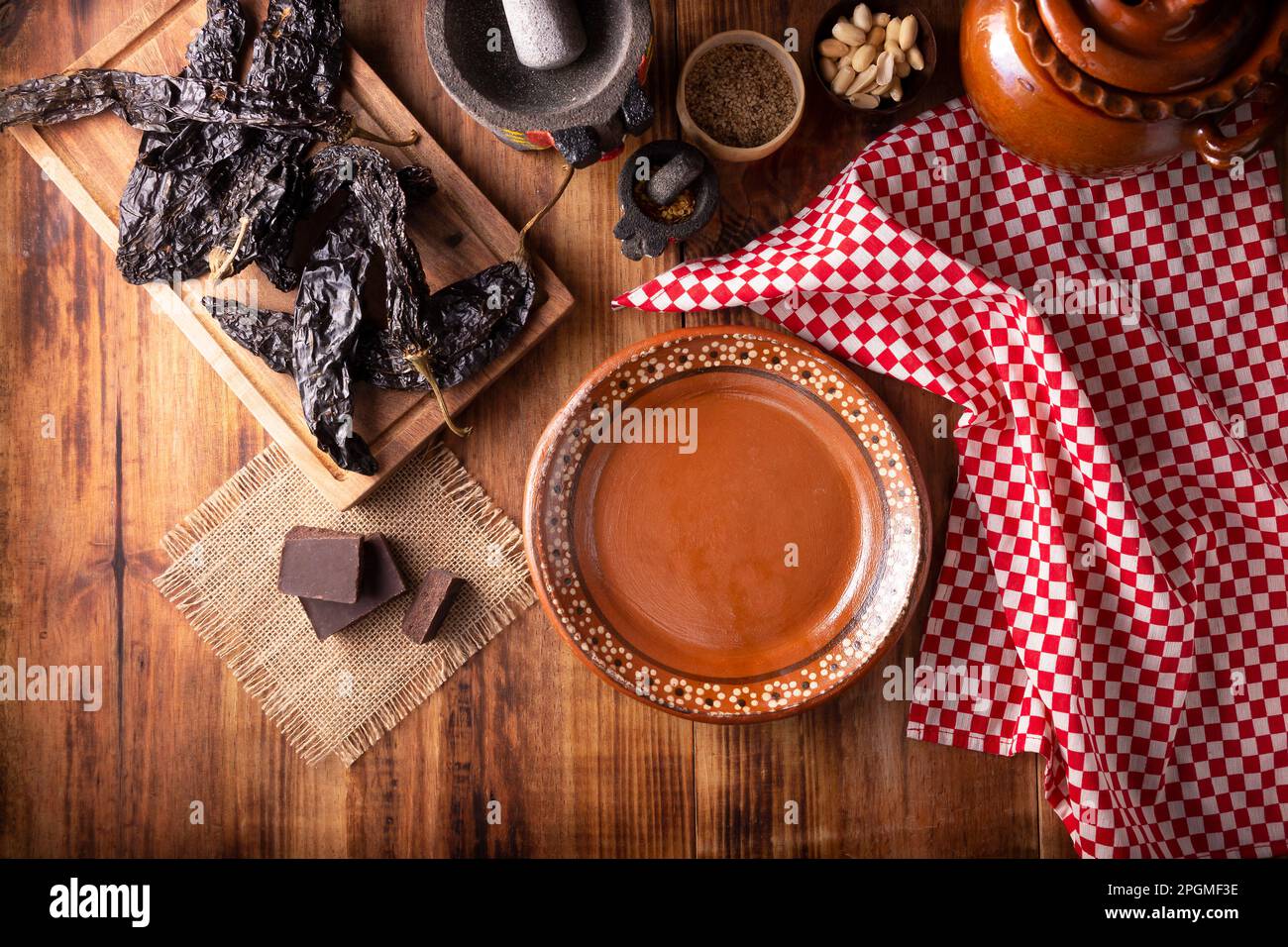 Leerer mexikanischer Tonteller mit einigen typischen Zutaten der mexikanischen Küche, Pasilla Chili, Schokolade, Molcajetes und Tontopf auf rustikaler Holzkarte Stockfoto