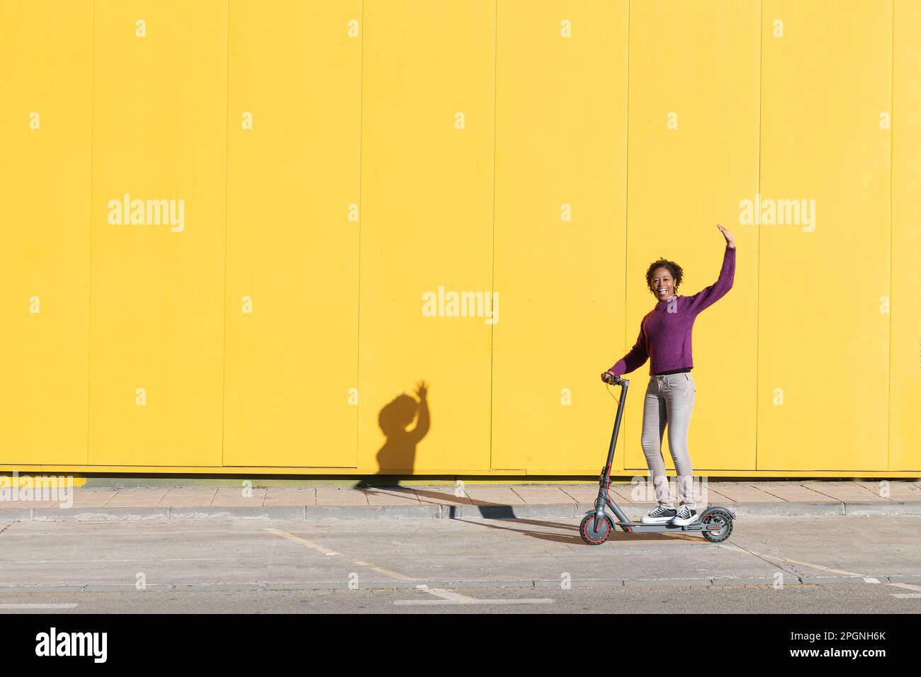 Glückliche Frau, die mit einem elektrischen Schubroller auf dem Fußweg winkt Stockfoto