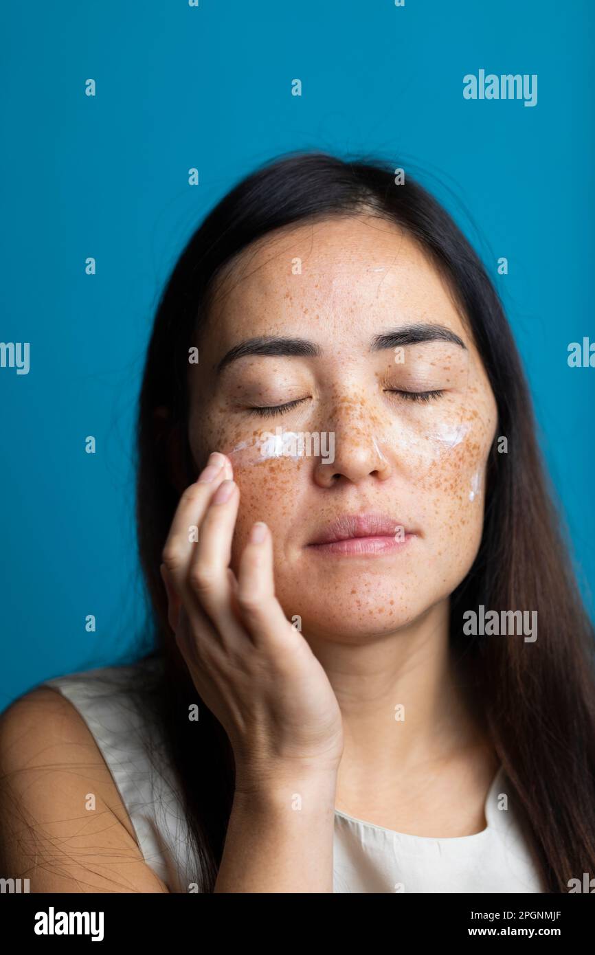 Frau mit geschlossenen Augen, die Feuchtigkeitscreme vor blauem Hintergrund aufträgt Stockfoto