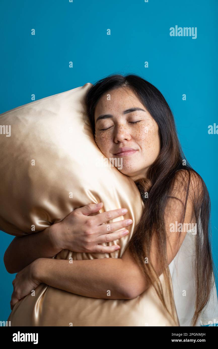 Frau mit geschlossenen Augen, die goldenes Kissen vor blauem Hintergrund umarmt Stockfoto