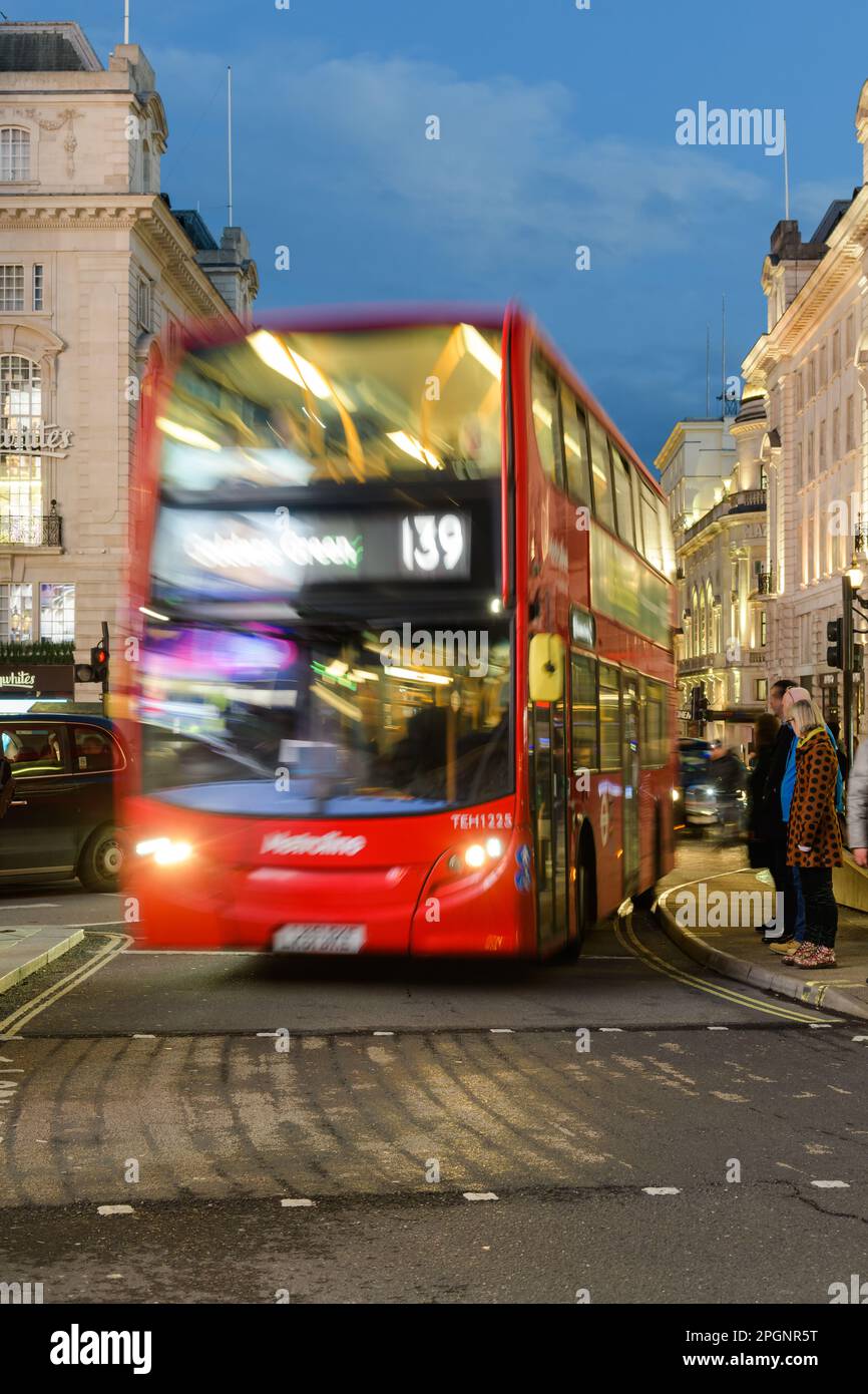 London, Großbritannien - 17. März 2023; verschwommener roter Londoner Bus fährt durch Piccadilly Circus mit Geschwindigkeit, während Fußgänger in der Abenddämmerung auf die Straße warten Stockfoto