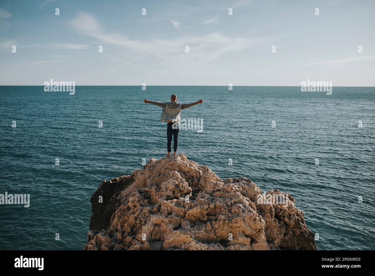 Ein sorgenfreier Mann, der mit gestreckten Armen vor dem Meer auf einem Felsen steht Stockfoto