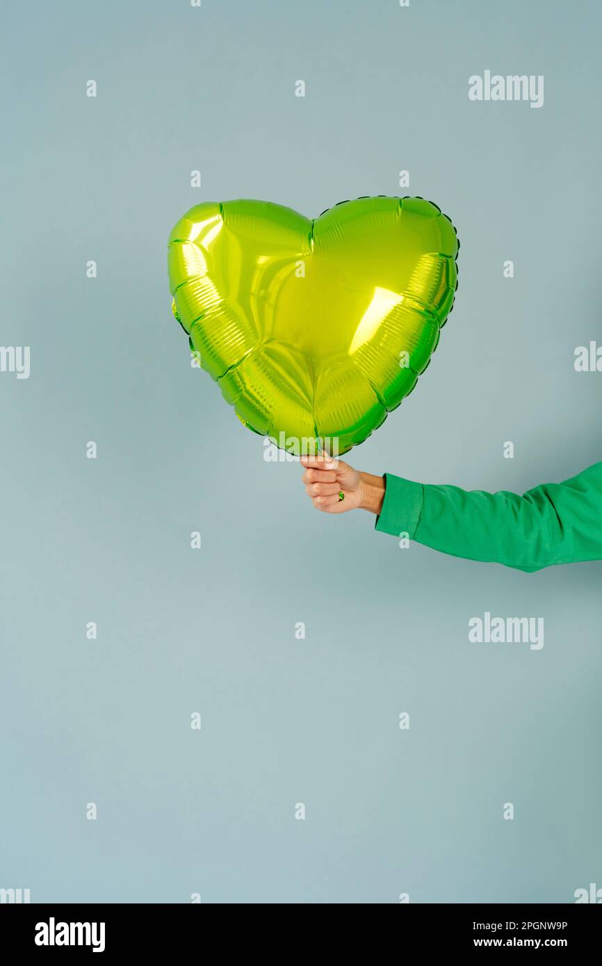 Eine Frau, die einen grünen, herzförmigen Ballon vor blauem Hintergrund hält Stockfoto