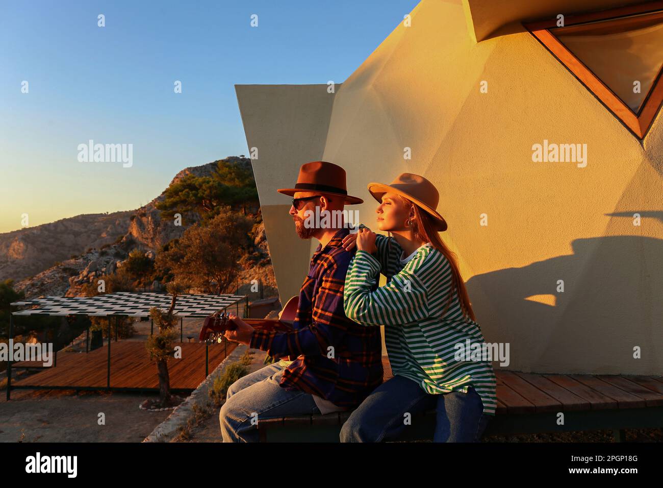 Ein Mann spielt Gitarre, sitzt mit einer Freundin in der Nähe des Öko-Hauses bei Sonnenuntergang Stockfoto