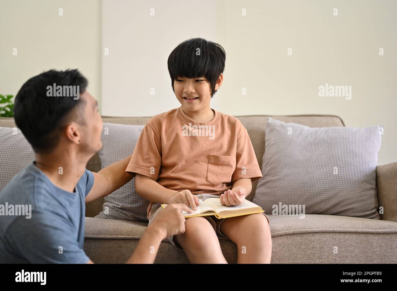 Ein bezaubernder und glücklicher junger asiatischer Junge, der mit seinem Vater spricht, während er Zeit verbringt und zusammen im Wohnzimmer ein Märchen liest. Alleinerziehender Vater und Kind Stockfoto