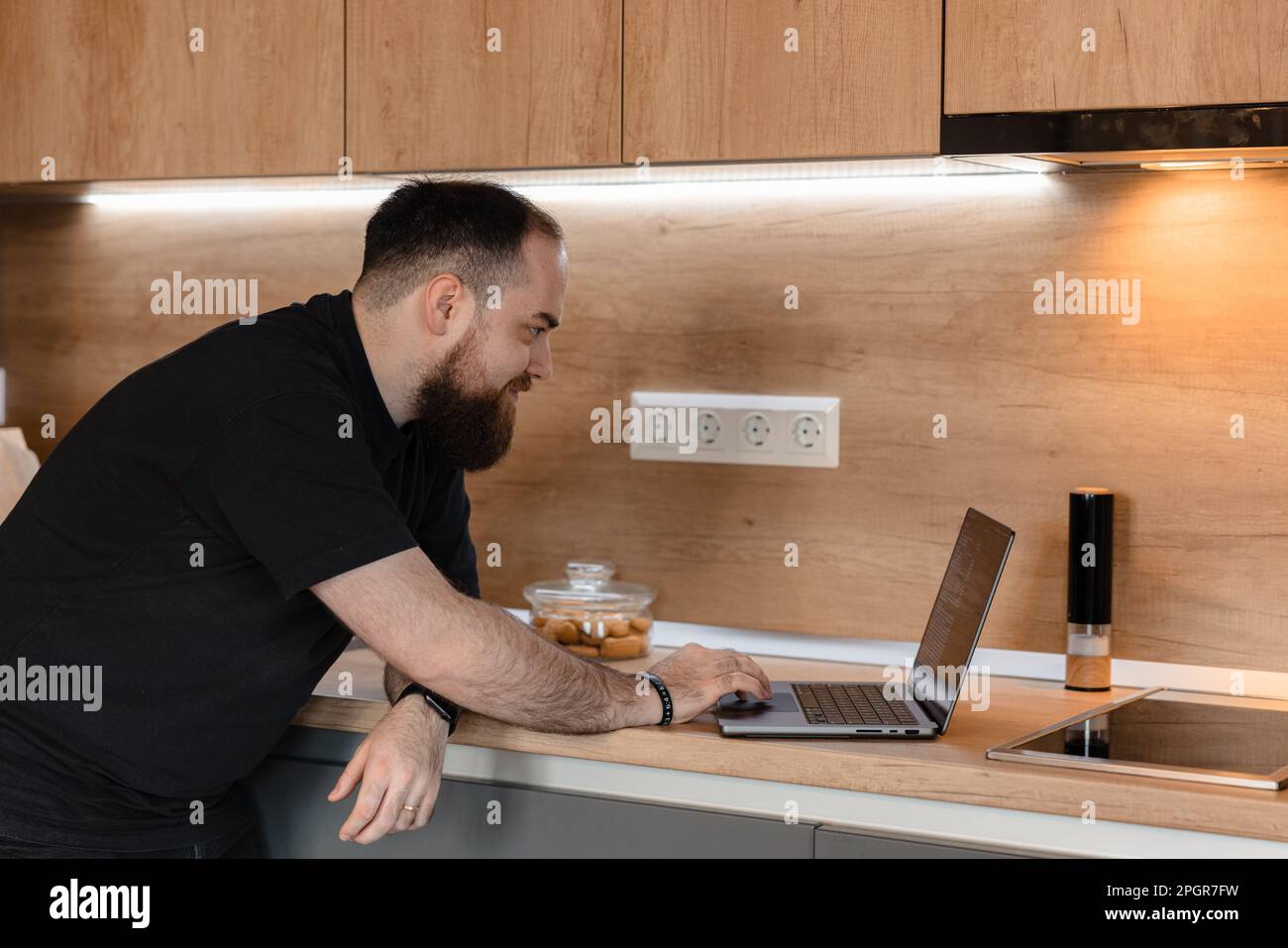 Ein Mann sitzt an seiner modernen Küchentheke und tippt auf seinem Laptop. Er trägt ein legeres T-Shirt und Jeans Stockfoto