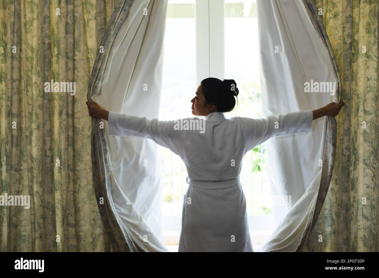 Glückliche Asiatin im Bademantel, die morgens die Vorhänge im Schlafzimmer öffnete. Häusliches Leben, Freizeit und Entspannung. Stockfoto