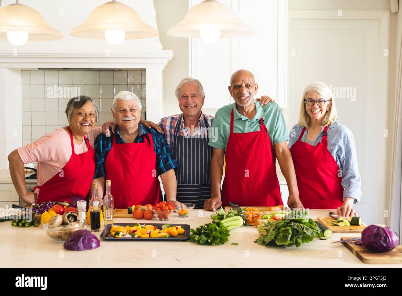 Porträt von glücklichen, vielfältigen Senior-Freunden, die Schürzen in der Küche tragen und sich während des Kochkurses umarmen. Ruhestand, Lernen, Kochen und gesundes Leben Stockfoto