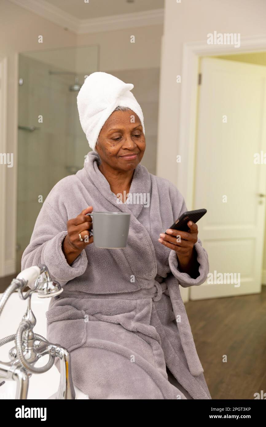 Eine afroamerikanische Seniorin, die eine Kaffeetasse in der Hand hält und ihr Handy benutzt, während sie in der Badewanne sitzt. Kopierraum, unverändert, Lebensstil, Baden, Trinken, ba Stockfoto