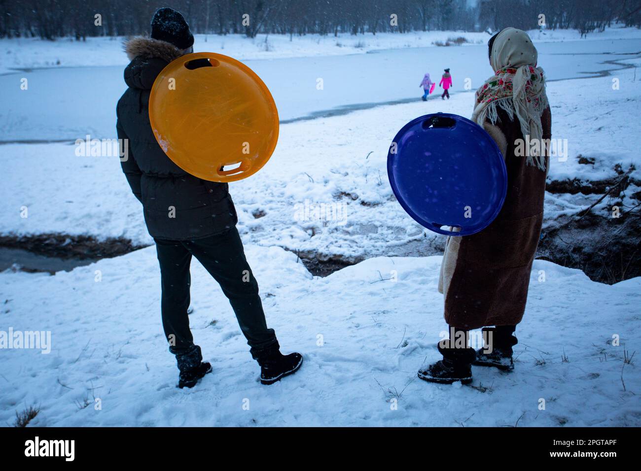 Friedliches Wochenende. Eltern in Kiew halten große Plastikteller, um im Schnee bergab zu fahren, beobachten ihre Kinder beim Spazierengehen am Ufer des gefrorenen Sees Stockfoto