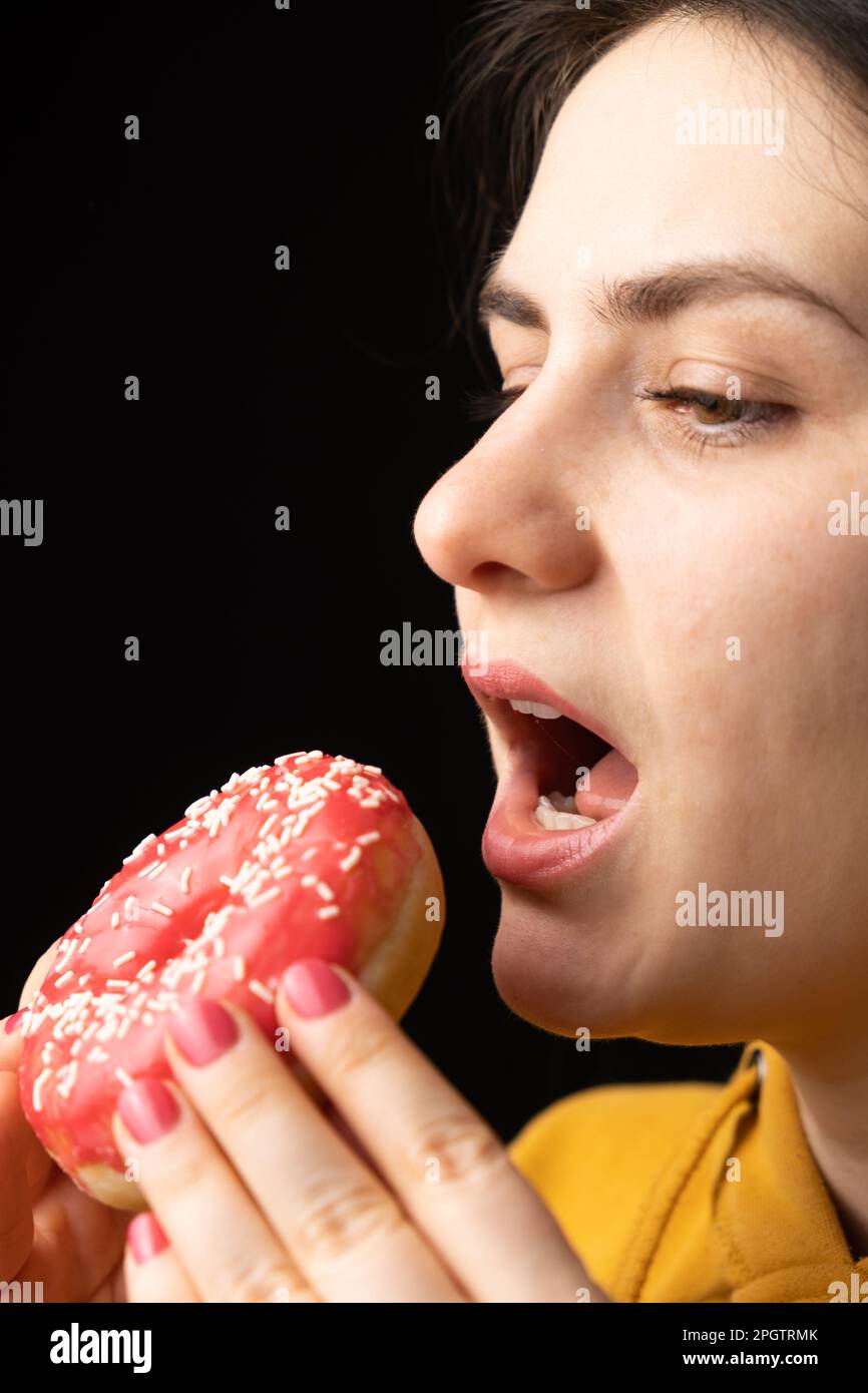 Eine Frau beißt einen großen roten Donut, einen schwarzen Hintergrund, ein Ort für Text. Gluttonie, überfressen und zuckersüchtig. Stockfoto