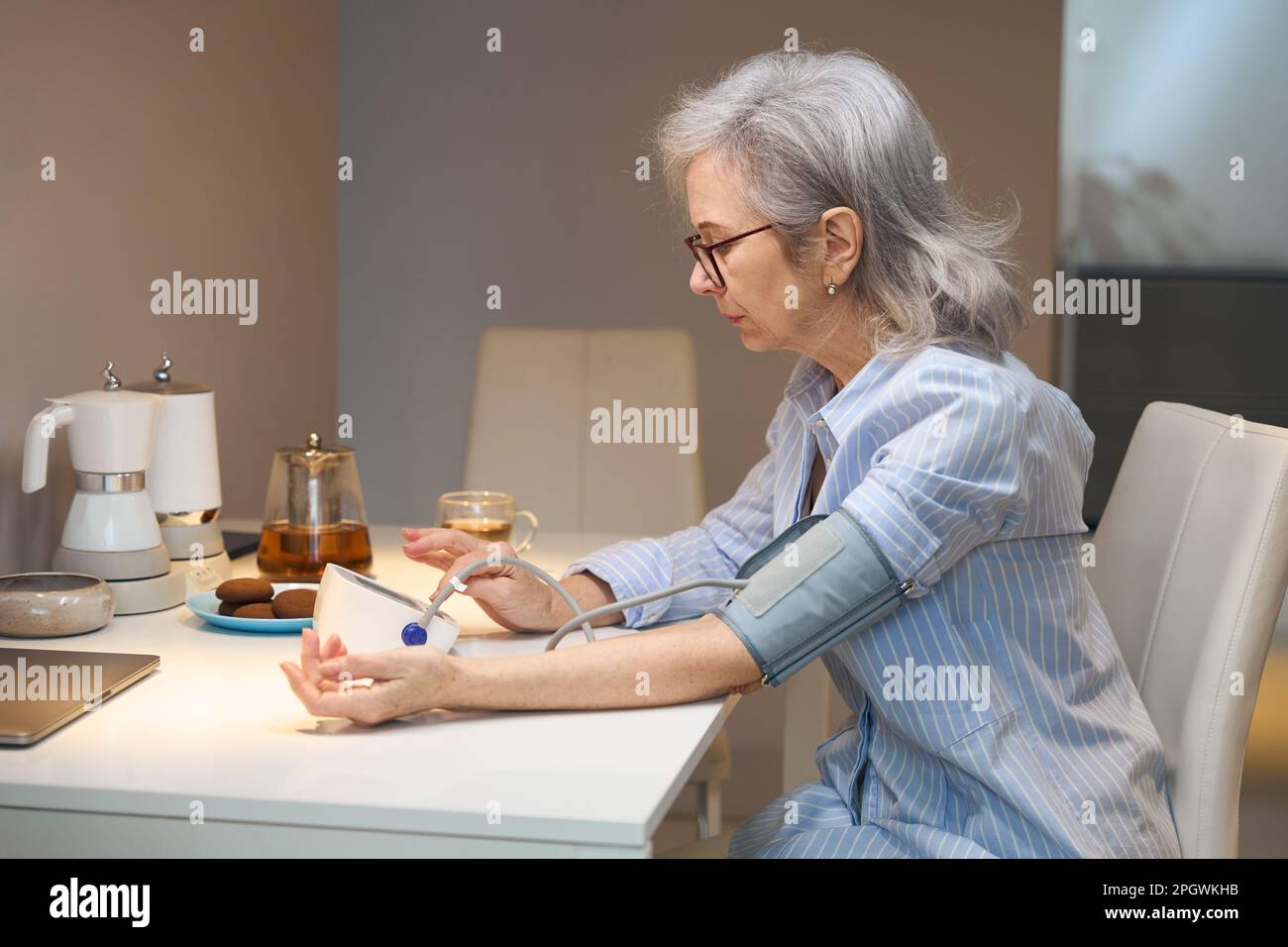 Die grauhaarige Dame sitzt am Küchentisch und misst ihren Blutdruck Stockfoto