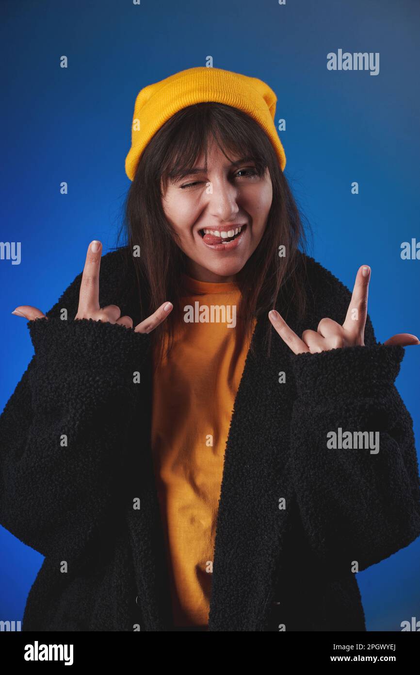 Vertikales Studioporträt einer jungen Frau in einem gelben Hemd, einer schwarzen Jacke und einer gelben Kappe, die mit beiden Händen, blinzelnd und hängend eine „Liebesgeste“ macht Stockfoto