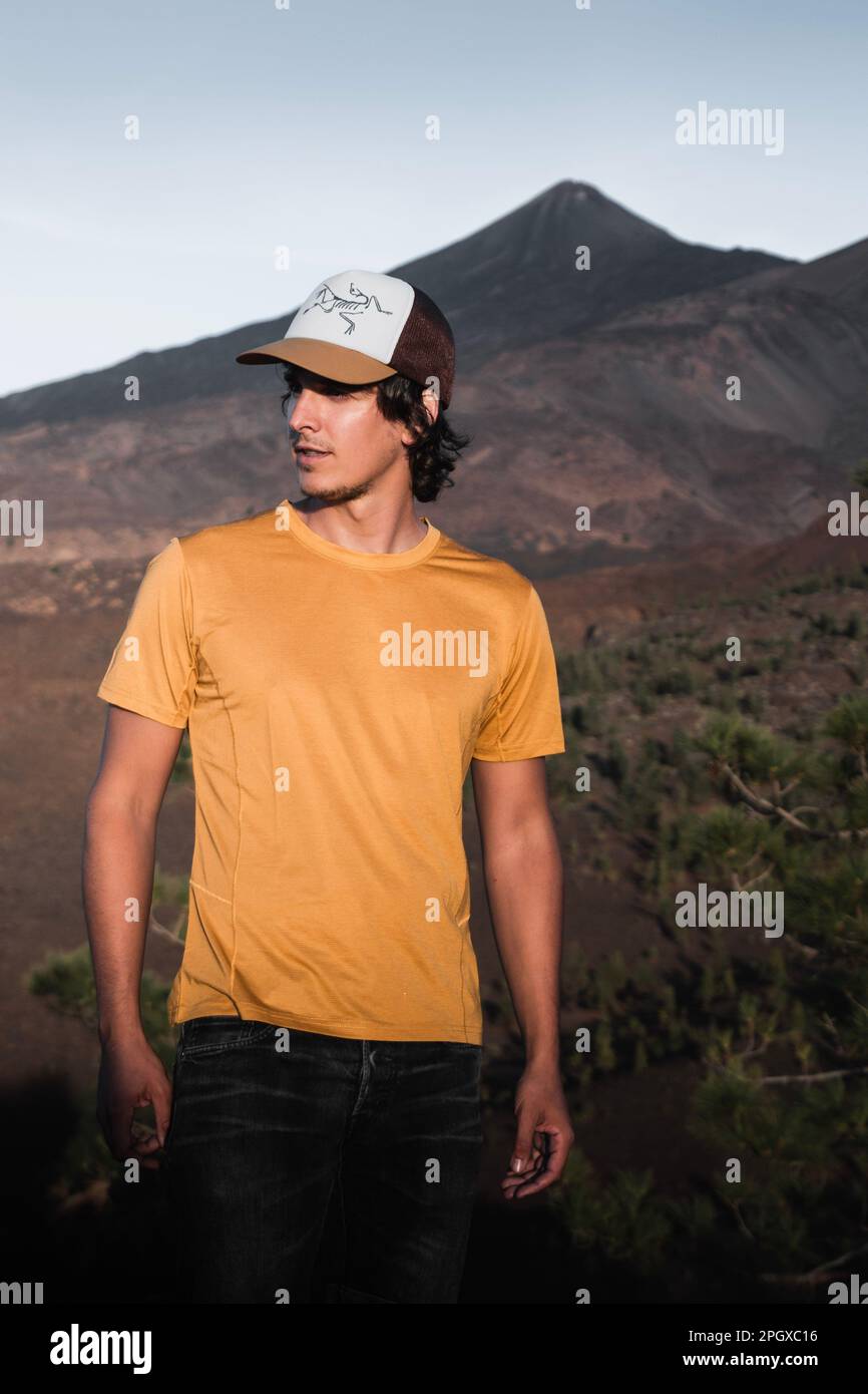 Junger Mann mit langen Haaren und einer Mütze mit gelbem Hemd in einer Berglandschaft bei Sonnenuntergang auf teneriffa. Stockfoto