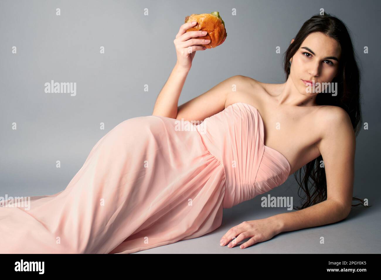 Eine Frau, die in einem durchgehenden pinkfarbenen Ballkleid mit einem Hamburger in der Hand lag und in die Kamera schaute Stockfoto