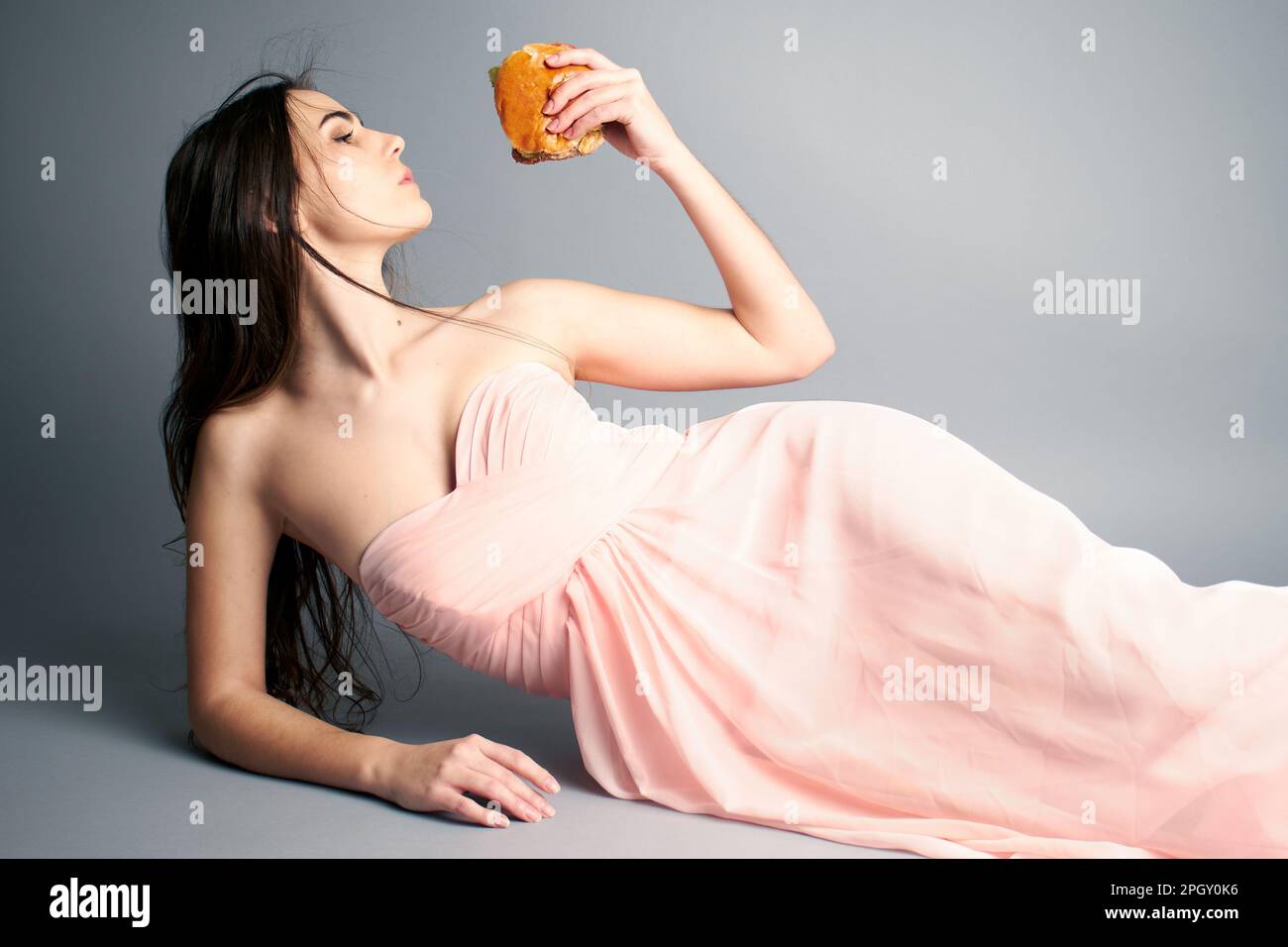 Porträt einer Frau, die einen Hamburger hält und Haare im Wind bläst, während sie sich im Ballkleid legt Stockfoto