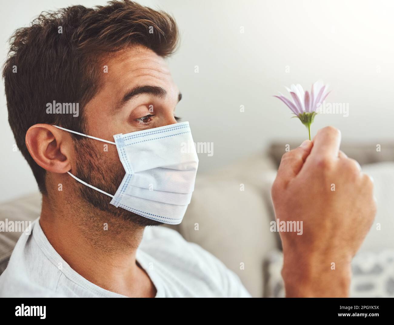 Kann ich überhaupt durch dieses Ding riechen? Ein attraktiver junger Mann, der eine Schutzmaske trägt, während er eine Blume hält. Stockfoto