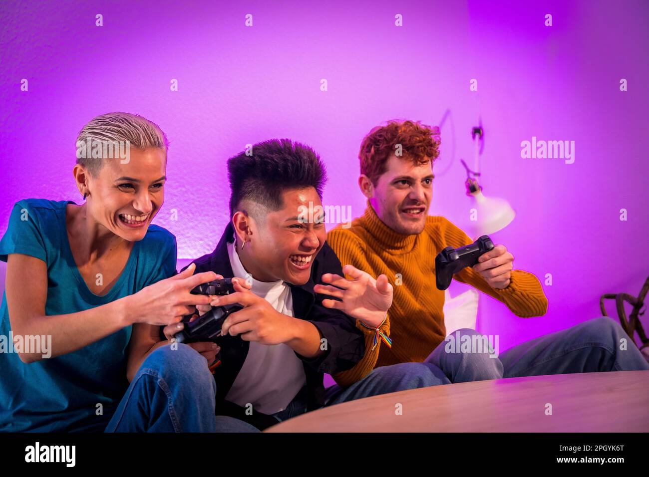 Eine Gruppe junger Freunde, die zusammen auf dem Sofa zu Hause Videospiele spielen, lila geführt, Spaß beim Schieben haben Stockfoto