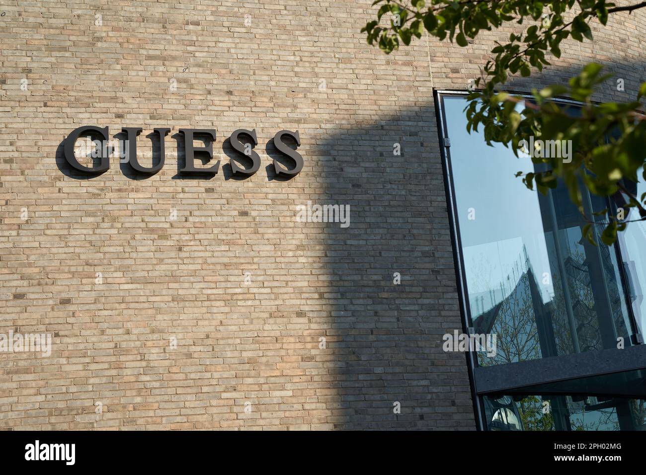 Metzingen - 03. Mai 2021: Guess Modefirma. Schwarze Buchstaben an grauer Ziegelwand. Shop-Fenster und grüne Zweige befinden sich auf der rechten Seite Stockfoto