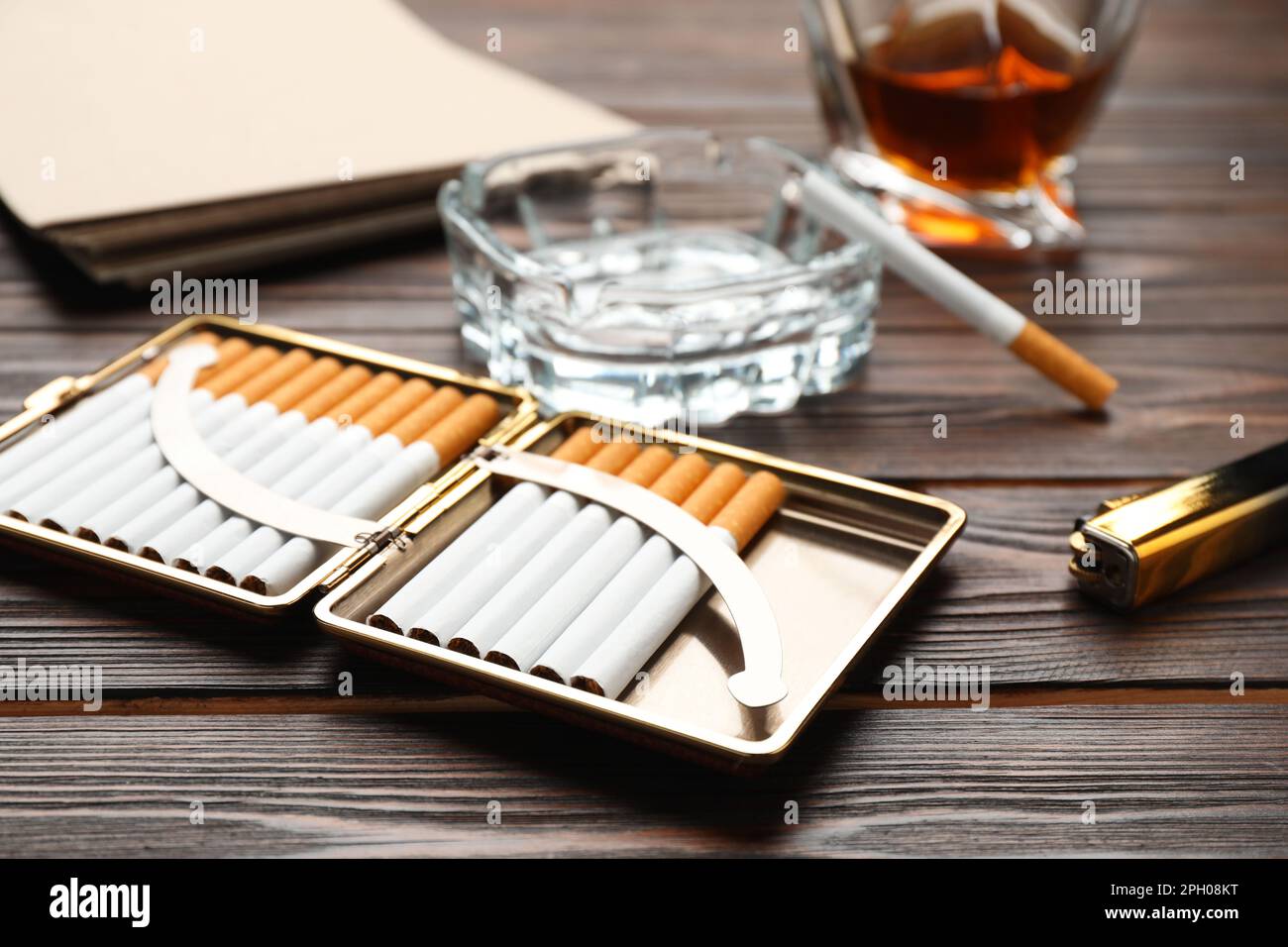 Offener Koffer mit Tabakfilterzigaretten, Aschenbecher, Alkoholgetränk und Feuerzeug auf Holztisch, Nahaufnahme Stockfoto