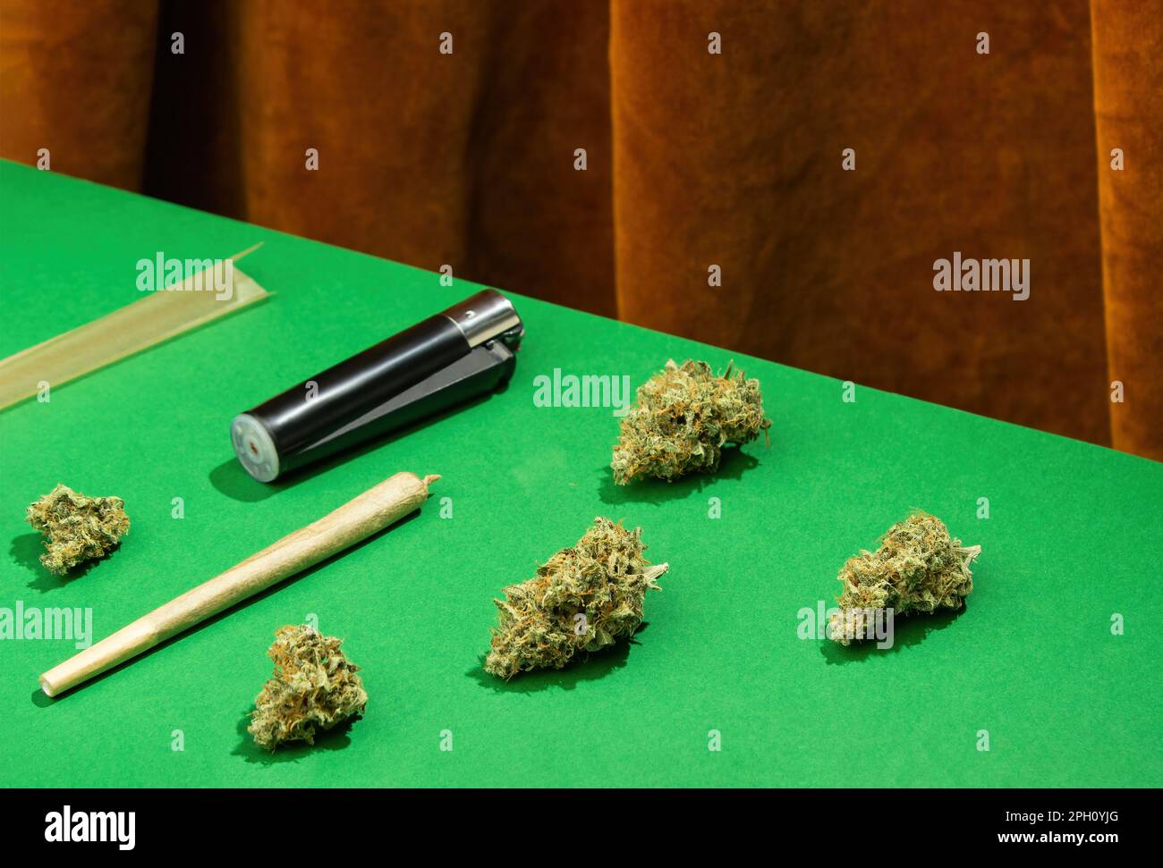 King-Size-Joint, Papier und Feuerzeug liegen zwischen trockenen Marihuana-Knospen auf einem grünen Tisch vor einem Hintergrund mit braunen Samtvorhängen Stockfoto