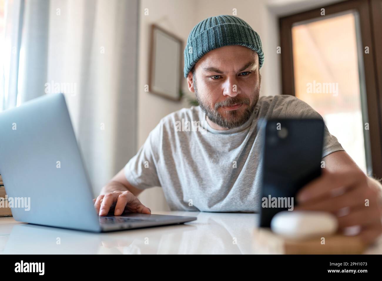Ein junger Erwachsener nutzt E-Banking auf seinem Smartphone, während er zu Hause mit einem Laptop arbeitet. Stockfoto