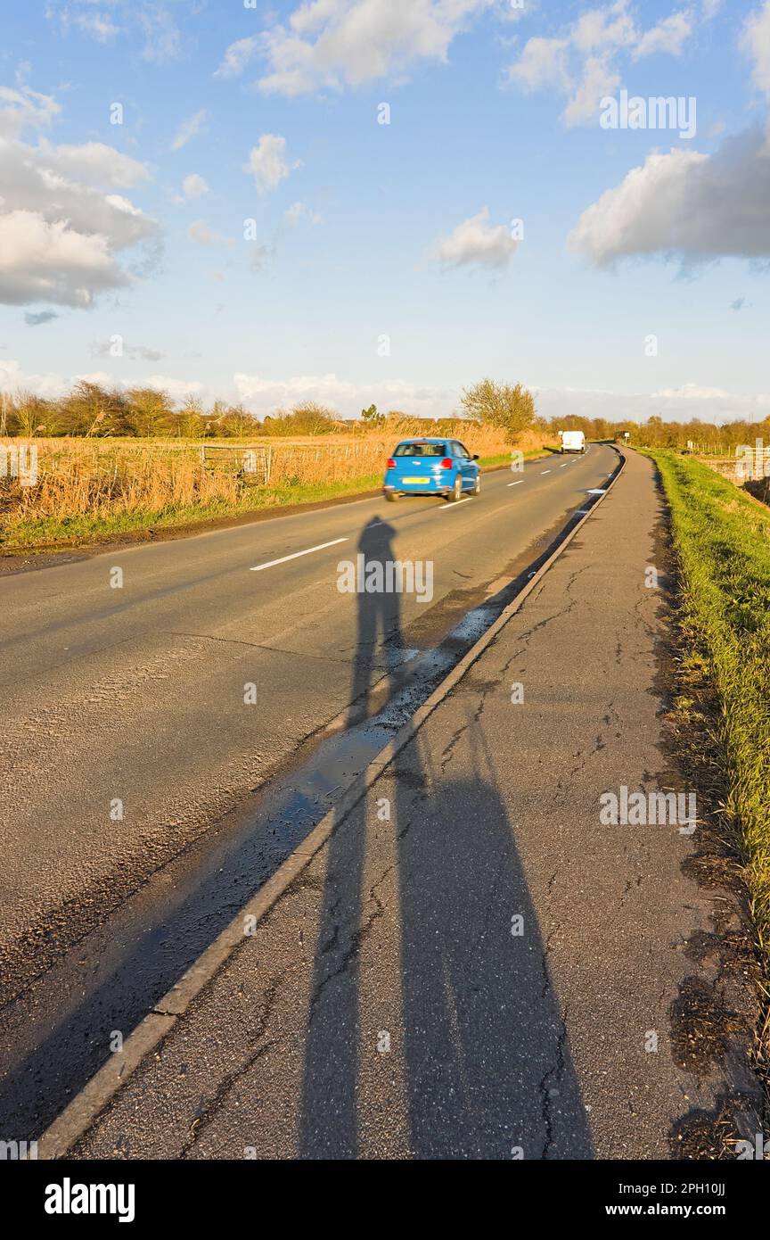 Schatten einer Person auf einer Landstraße mit einem Auto, das vorbeifährt und in die Ferne fährt Stockfoto