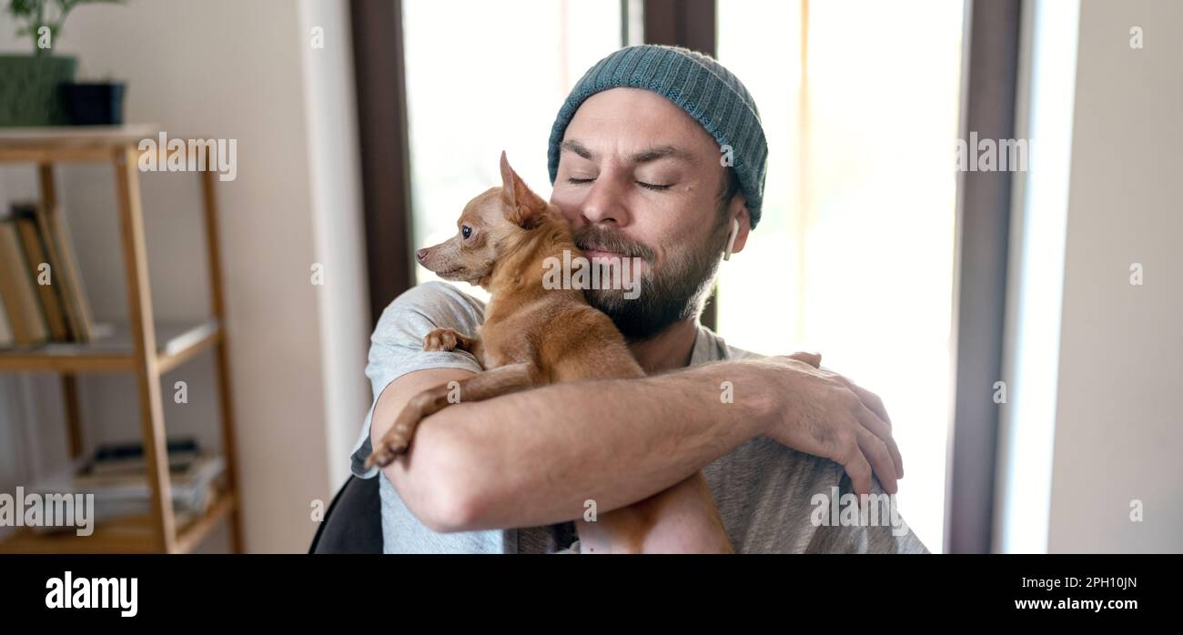 Ein erwachsener bärtiger Mann mit Hut umarmt seinen kleinen Hund, seine Liebe und Zärtlichkeit. Stockfoto