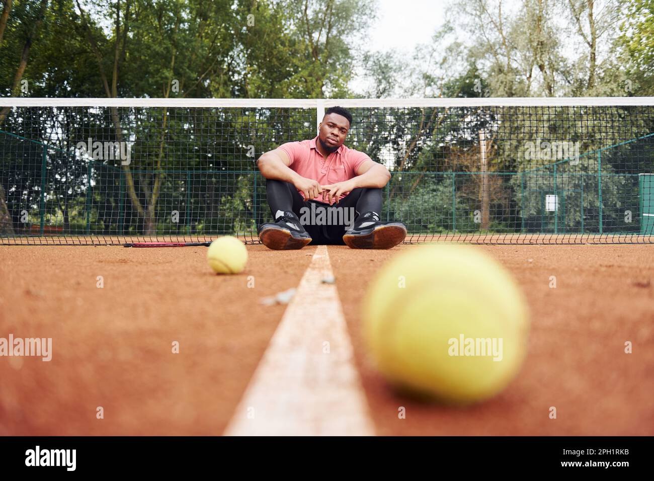 Er sitzt in der Nähe des Netzes und macht eine Pause. Ein afroamerikanischer Mann in einem rosa Hemd sitzt mit einem Tennisschläger draußen auf dem Platz Stockfoto