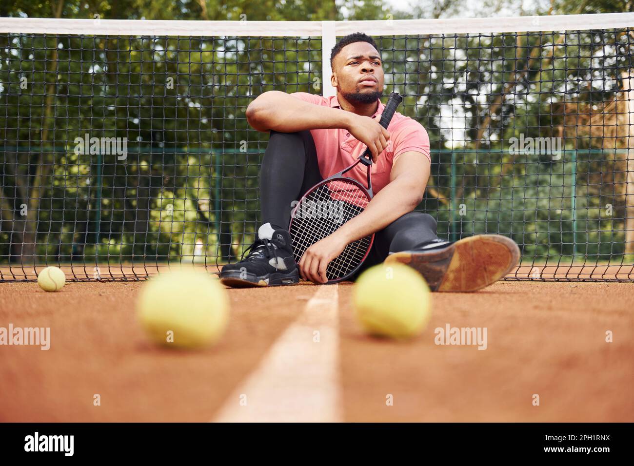 Er sitzt in der Nähe des Netzes und macht eine Pause. Ein afroamerikanischer Mann in einem rosa Hemd sitzt mit einem Tennisschläger draußen auf dem Platz Stockfoto