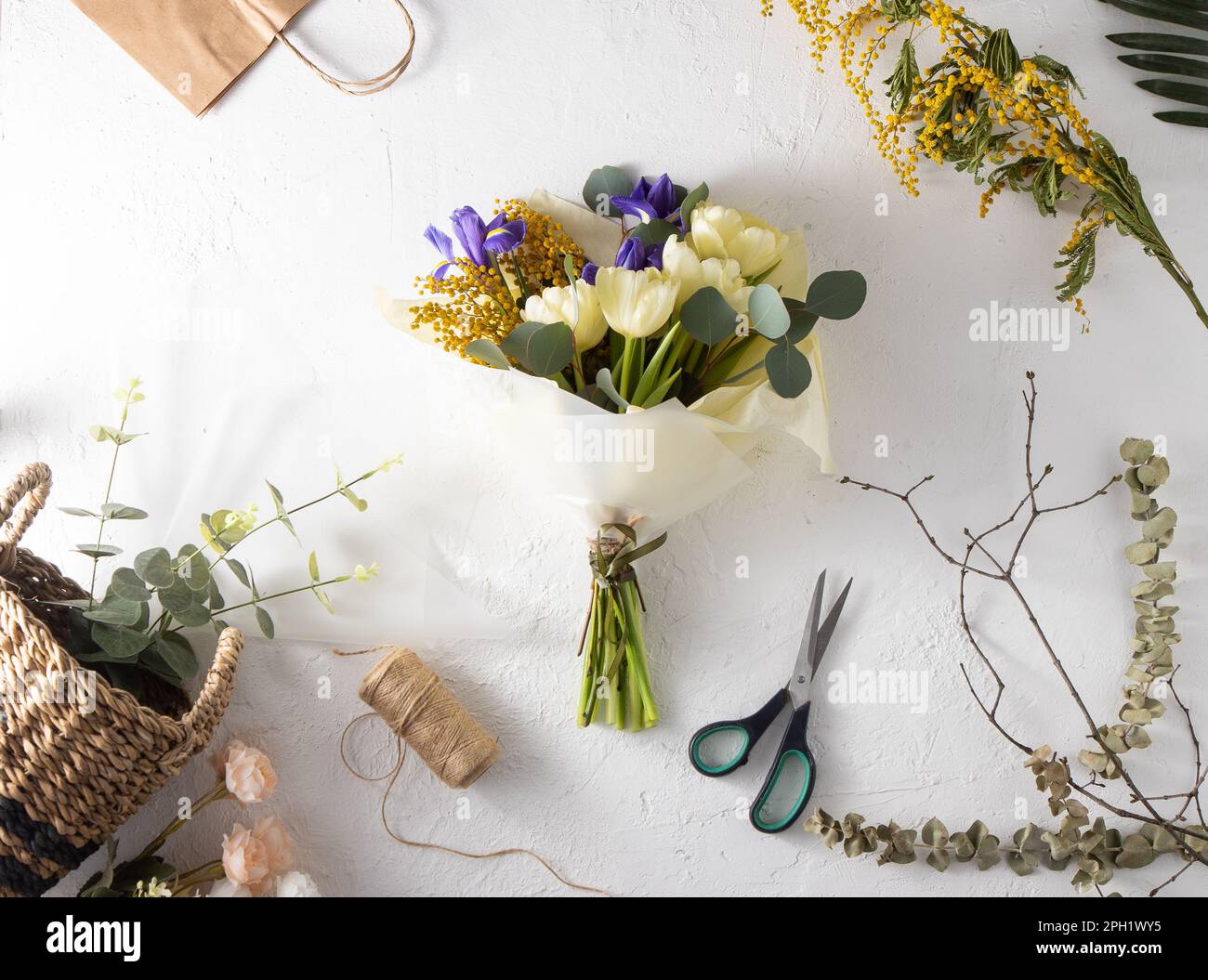 Machen Sie Ihren Blumenstrauß zum Arbeitsplatz. Blumenarrangements in einem Blumenladen. Frühlingsstrauß gelber Blumen. Iris, Tulpen, Mimosa und Eukalyptus. Tulpenknospe. Blumenhintergrund. Lila Iris. Weißer Tisch Stockfoto